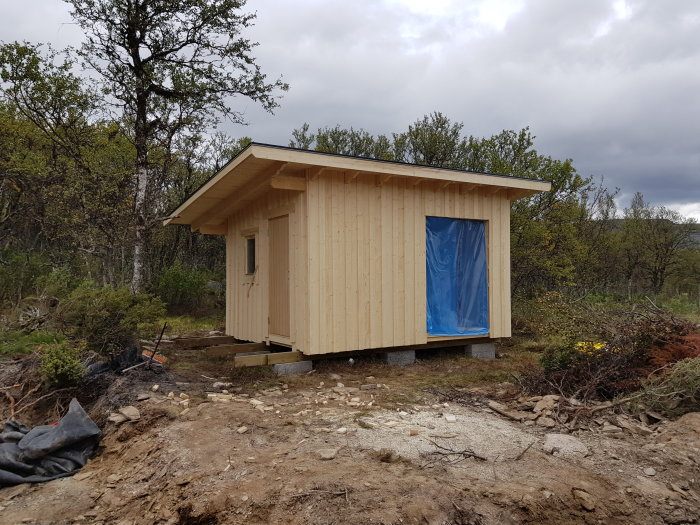 Nybyggt träskjul på landsbygden med blå presenning framför ingången och natur i bakgrunden.