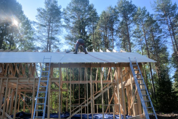 Arbetare i säkerhetssele fäster takplattor på en husstomme med en röd säkerhetslina synlig.