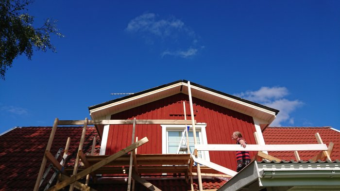 Person som arbetar på ett rött hus med nyinstallerade vindskivor och trekantslister under en klarblå himmel.