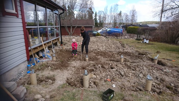 Schaktad trädgård med betongplintar för altanbygge, vuxen och barn granskar arbetet.