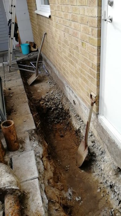Grävningsarbete längs husgrund för installation av ny dränering med spade och grushög.