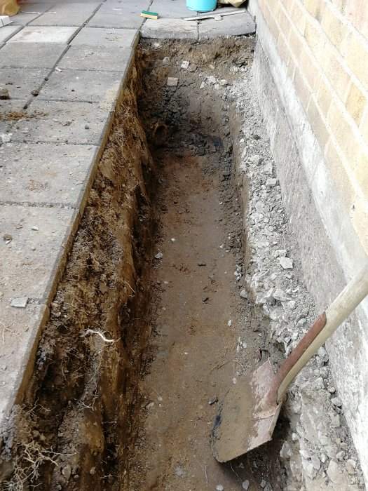 Grävd schakt i trädgård intill tegelvägg med spade, klar arbetsdag.