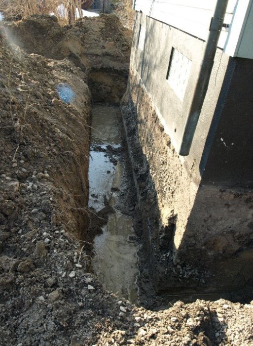 Grävd grund vid husvägg med stora vattenpölar och lös jord.