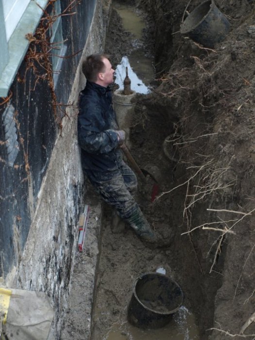 Person gräver i en smal och djup grävning nära en husvägg, lerig och utmanande arbetsförhållande.