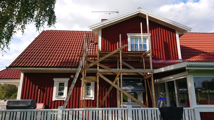 Ett rött hus med en demonterad ställning framför ett delvis reparerat altantak, med en stege lutad mot väggen.