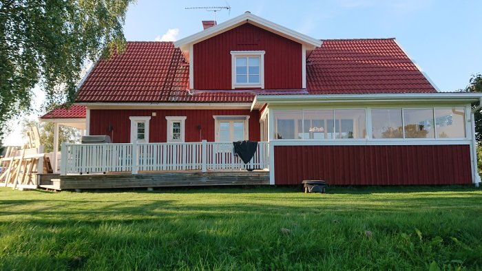 Rött hus med nyrenoverad veranda och plasttäckt fönsterparti, omgiven av en grön gräsmatta.