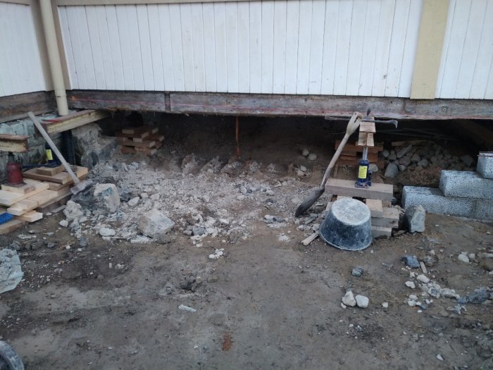 Husgrund med synliga stenar och grus under renovering, tillsammans med byggverktyg och stödben.