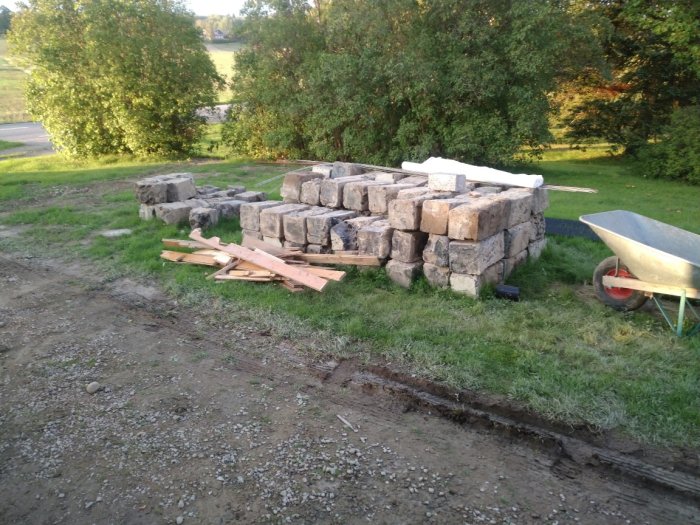 Stapel med stora flyttade stenar i en trädgård vid sidan av en skottkärra och timmer.
