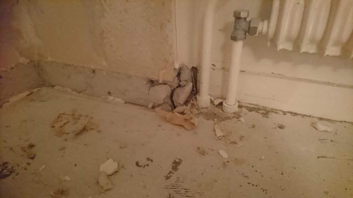 Skadad lättbetong vid golvhörn bredvid vitt rör och under radiator, med borttagen sockel och spill.