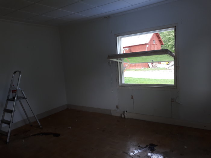 Rum under renovering med borttagen kakelugn, tomma väggar och trägolv, stege till vänster, fönster med utsikt över gård.