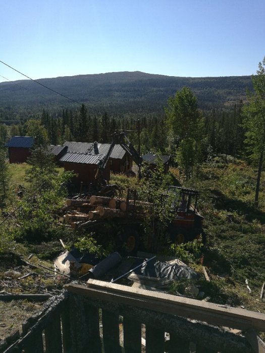 Skogsmaskin skotar stockar nära en stuga med berg i bakgrunden.