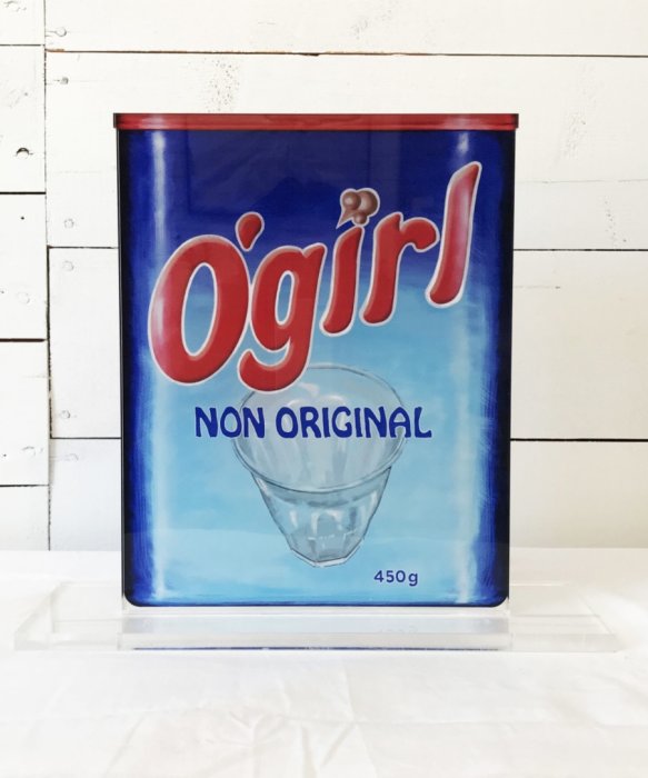 Blå förpackning med texten 'Ögirl NON ORIGINAL' ovanför en ritad glasskål mot en vit träbakgrund.