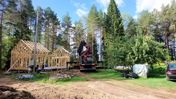 Kranbil vid byggande av trästomme för hus, omgiven av skog.