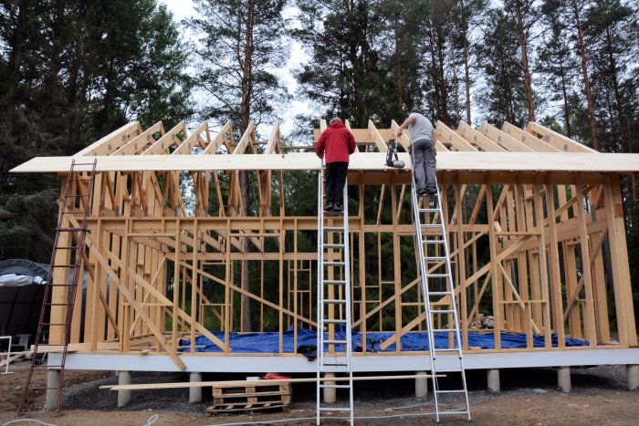 Två personer jobbar på taket av en pågående trähuskonstruktion med synligt råspont och stegar.