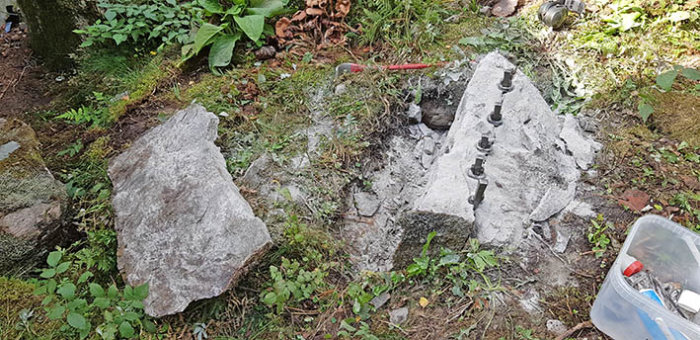 Stenblock i en trädgård delvis uppdelat med en stålspett och borrhål, omgiven av grönska och verktyg.