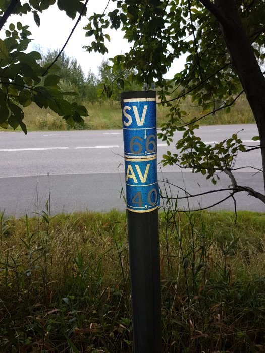 Stolpe med texten SV 6,6 och AV 4,0 vid en väg med grönska i bakgrunden.