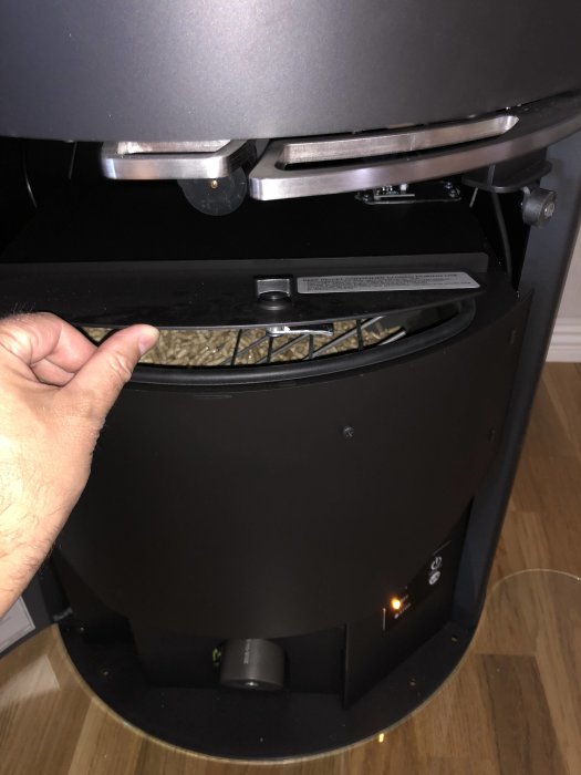 Hand öppnar luckan på ett svart pelletsförråd till en kamin, synligt fylld med träpellets, inget nivåfönster.