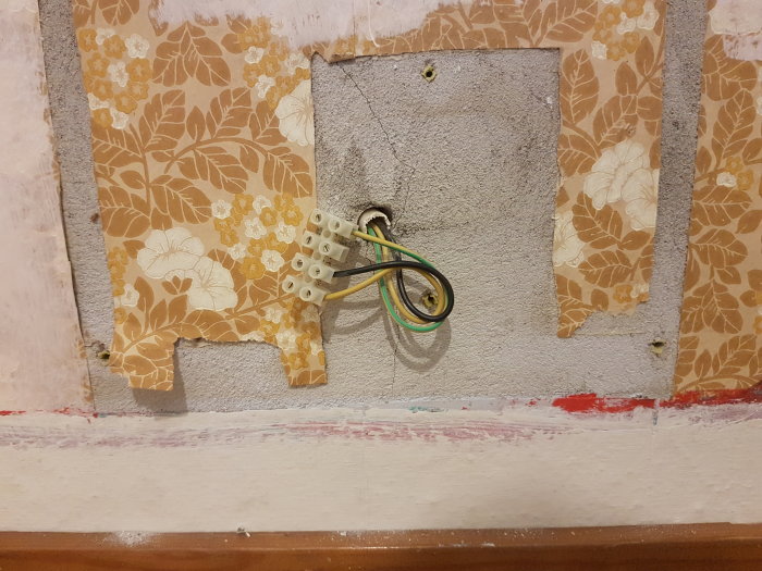 Elektriska ledningar sticker ut från en öppen vägg utanför en gammal radiatorplats med retro tapet.