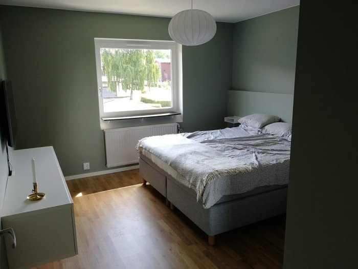 Renoverat sovrum med parkettgolv, grå säng och gröna väggar samt ett öppet fönster.