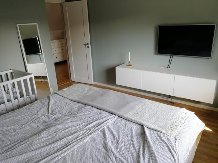 Renoverat sovrum med spjälsäng, dubbelsäng, vit innerdörr, TV och vit byrå mot en grön vägg.