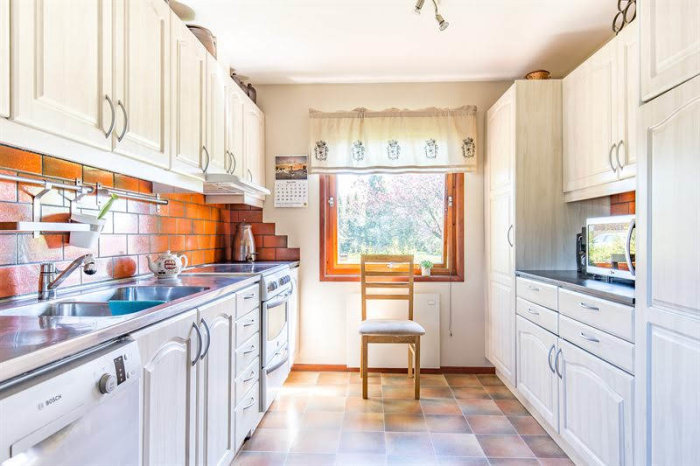 Retrokök med vita skåp, tegelröd kakelvägg, ljus inredning och fönster med utsikt mot trädgården.