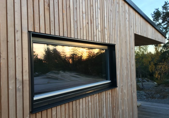 Stående träpanel med rak fals på husfasad med ett fönster och skymt av skog i bakgrunden.
