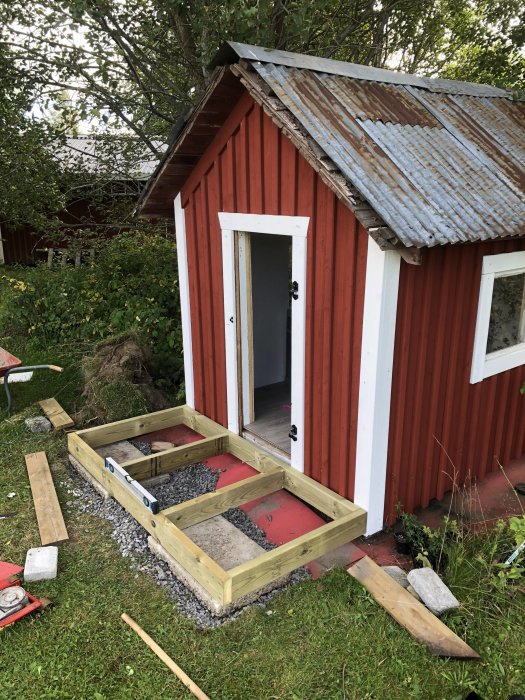 En röd lekstuga under renovering med nybyggd träram i förgrunden och verktyg utspridda på marken.