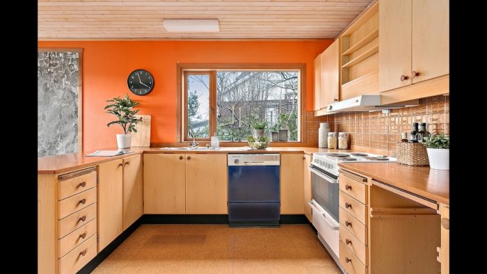 Kök med träskåp, orangea väggar och brunt kakel ovan diskbänk, inredning känns daterad.