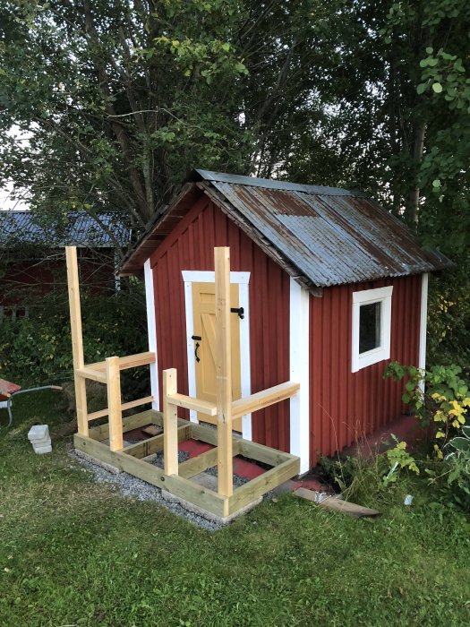 Halvfärdig trälekstuga med röda väggar och slitet plåttak, påbörjad trätrappa framför.