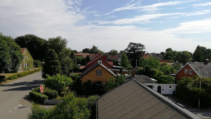 Utsikt över bostadsområde med tegeltak och gröna träd mot en blå himmel med ljusa moln.