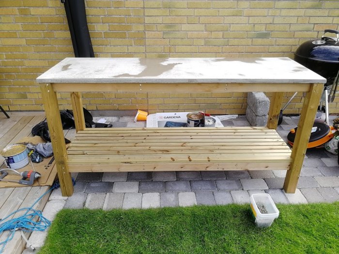 Nybyggt trägrillbord framför en gul tegelvägg omgiven av byggmaterial och grillutrustning.