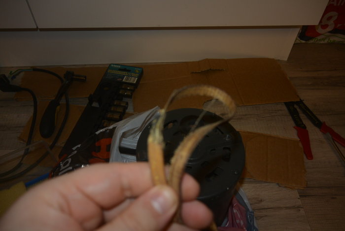 Hand håller ett slitet sågblad framför en samling verktyg och byggmaterial på golvet.