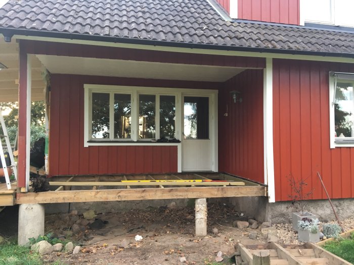 Rødmalet hus med træveranda under ombygning, mangler en bærende stolpe, set foran gruset fundament og haveindretning.