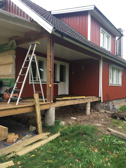 Rødmalet hus med en halvfærdig terrasse, stige op ad væggen. En plint uden pelare, byggeudstyr og træmaterialer ligger på græsset.