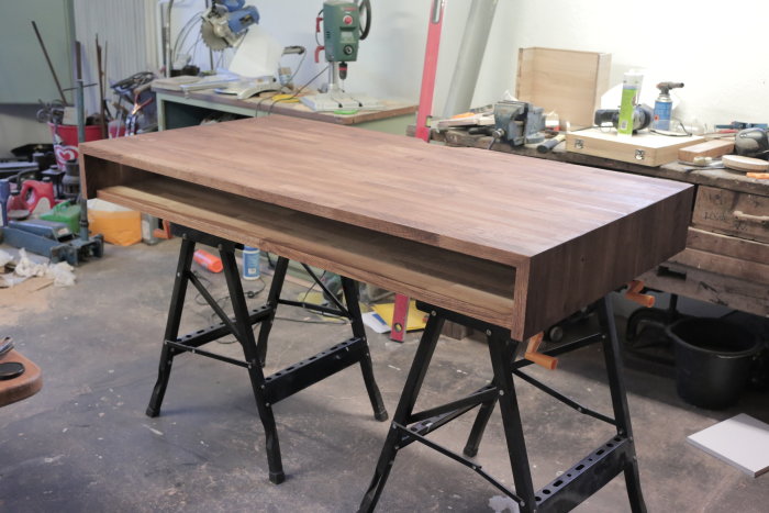 Träs skrivbord på sågbockar i en verkstad, lim och sågspån syns i en glipa.