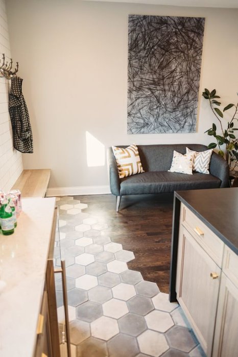 Hexagonalt klinkergolv i övergång till mörkt trägolv i ett modernt vardagsrum med grå soffa och konstverk.