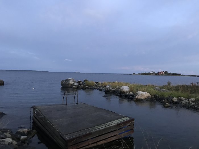 Kvällsutsikt över sjö med brygga i förgrunden, ö till vänster och fartyget TRANSLONTANO på ankare i fjärran.