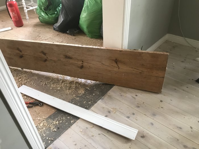 Ett gammalt upptaget plankgolv med synliga spikhål och märken, omgiven av sågspån och renoveringsmaterial.