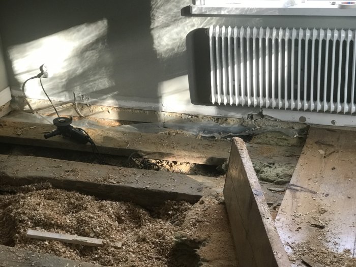 Upprivet gammalt plankgolv med synligt isoleringsmaterial och verktyg vid en vägg.