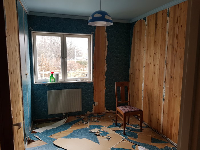 Rum under renovering, blå blommig tapet delvis avriven, plastmatta på golvet och oisolerade väggar.