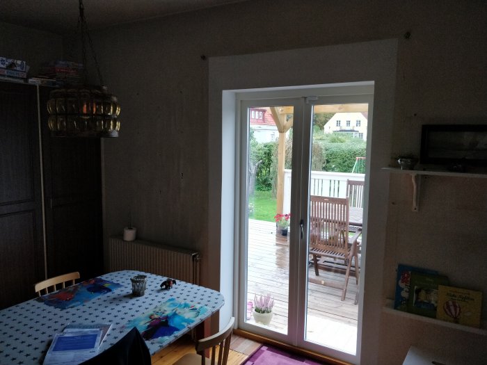 Skyvedør til terrasse fra kjøkken, med bord og stoler på utsiden. Inne er et spisebord med duk og barnerelatert dekor. Vegg skal åpnes opp.