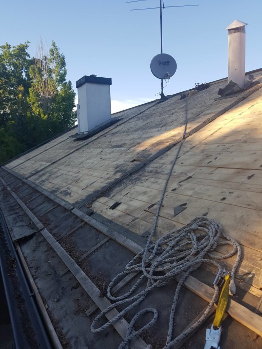 Tak under ombyggnad med ny spont, rep och arbetsverktyg synligt, samt skorstenar och parabolantenn i bakgrunden.