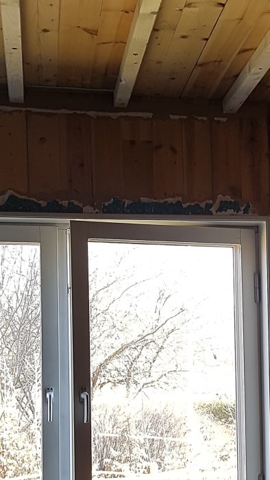 Fönster med synlig övre del och takbjälkar, fogskum syns mellan fönster och träpanel ovanför.