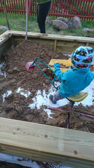 Barn i hjälm leker med skottkärra och murarsand vid en nybyggd sandlåda.