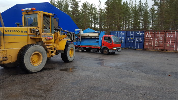Gul Volvo grävmaskin, blå lastbil och röd bil framför staplade fraktcontainrar på en grusplan.
