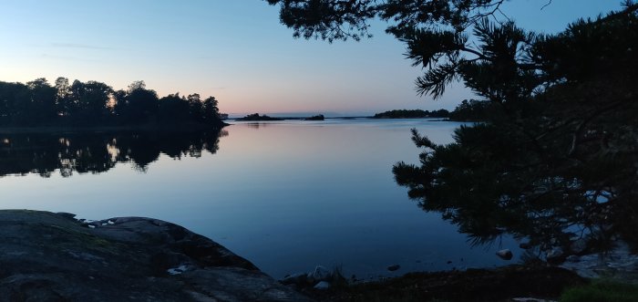 Solnedgång över en stilla skärgårdsvik med träd och klippor i förgrunden.