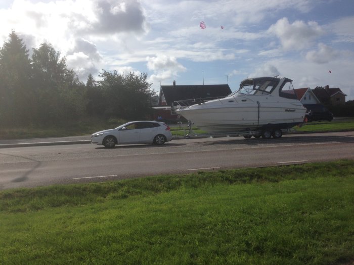 Personbil som drar en stor båt på en trailer längs en väg, med hus och grönska i bakgrunden.