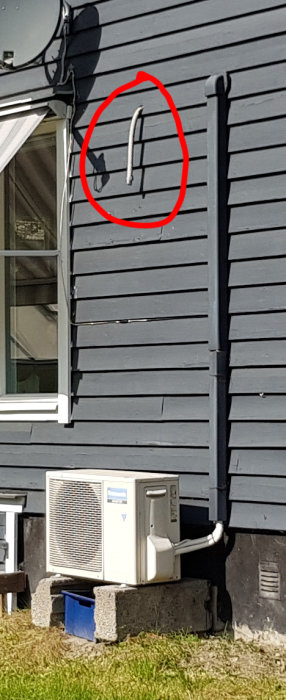 Luftvärmepump installerad vid husvägg med markerat rör på fasaden, misstänkt var ett dräneringsrör.