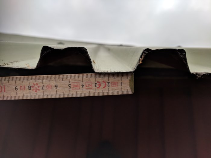 Mått av plåtprofil med tumstock på en skadad takdel för identifiering av plåttyp.