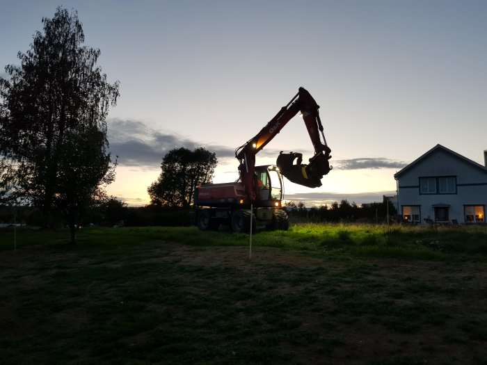 Grävmaskin och lastbil vid solnedgång i trädgård nära ett hus, kanske början på ett byggprojekt.